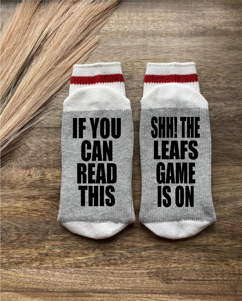 May include: A pair of gray socks with white and red stripes. One sock says "If you can read this" and the other sock says "Shh! The Leafs game is on".