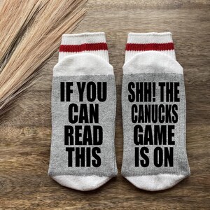 May include: A pair of gray crew socks with white and red stripes at the top. One sock says "If you can read this" and the other sock says "Shh! The Canucks game is on".