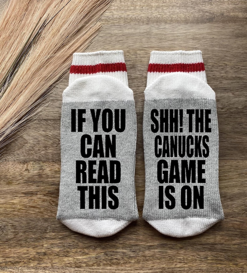 May include: A pair of gray crew socks with white and red stripes at the top. One sock says "If you can read this" and the other sock says "Shh! The Canucks game is on".