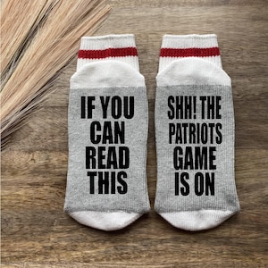May include: A pair of gray and white socks with red stripes. One sock says "If you can read this" and the other sock says "Shh! The Patriots game is on".