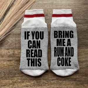 May include: A pair of gray socks with red and white stripes at the top. One sock says "If you can read this" and the other sock says "Bring me a rum and coke".