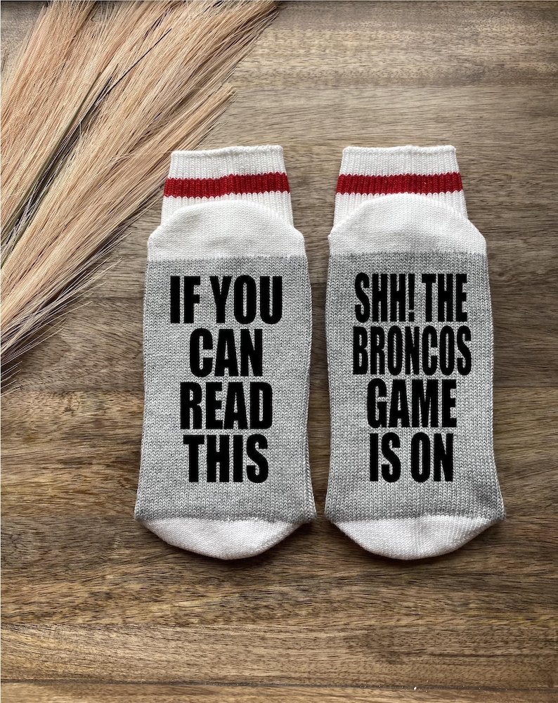 May include: A pair of gray socks with red and white stripes at the top. One sock says "If you can read this" and the other sock says "Shh! The Broncos game is on".