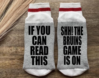 May include: A pair of gray socks with red stripes at the top. One sock says "If you can read this" and the other sock says "Shh! The Bruins game is on".