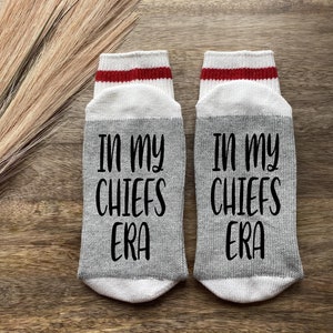 K&ouml;nnte beinhalten: Ein Paar graue Socken mit wei&szlig;en und roten Streifen oben. Die Socken haben den Text "In my Chiefs Era" in Schwarz aufgedruckt.