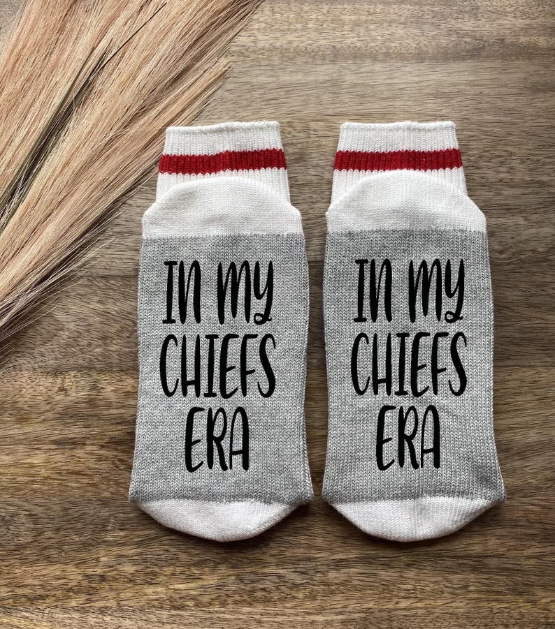 K&ouml;nnte beinhalten: Ein Paar graue Socken mit wei&szlig;en und roten Streifen oben. Die Socken haben den Text "In my Chiefs Era" in Schwarz aufgedruckt.