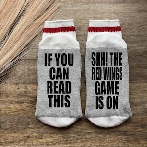 May include: A pair of gray socks with white and red stripes at the top. One sock says "If you can read this" and the other sock says "Shh! The Red Wings game is on".