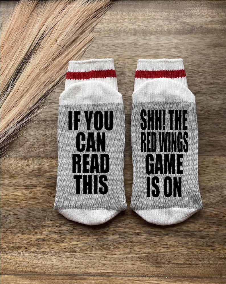 May include: A pair of gray socks with white and red stripes at the top. One sock says "If you can read this" and the other sock says "Shh! The Red Wings game is on".