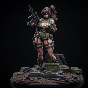 Op de afbeelding: Gedetailleerd beeldje van een vrouwelijk personage in militaire kleding, met een geweer. Het beeldje staat op een diorama-basis met rotsen en kleine kisten. Het kleurenpalet omvat tinten groen, bruin en zwart.