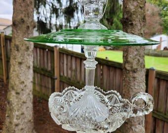 May include: A hanging bird feeder constructed from clear and green pressed glass. The structure features a green glass plate top, a clear glass pedestal stem, and a clear glass bowl base with a decorative handle, all suspended by a wire.