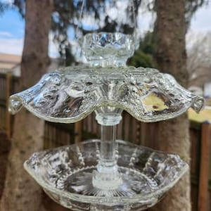 May include: A clear glass bird feeder with a tiered design. The feeder has a round base, a central support, and a decorative top with a candle holder. The glass has embossed floral patterns. It is hanging outdoors.