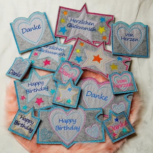 Peut inclure: Une collection d'appliques en feutre avec différents motifs et phrases, notamment "Happy Birthday", "Danke" et "Von Herzen". Les appliques sont dans des tons de gris, bleu, rose et jaune.