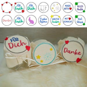 Op de afbeelding: Drie glazen potten met stoffen deksels. De deksels zijn versierd met geborduurde ontwerpen. Een deksel heeft een sneeuwvlok en sterren, een ander heeft de tekst "Danke" en een hart, en de derde heeft de tekst "FÜR Dich" en een hart.