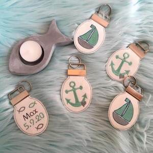 Pode incluir: Cinco chaveiros com designs diferentes. Cada chaveiro é um oval de cor creme com um design bordado em verde. Os designs incluem um veleiro, uma âncora e um peixe. Um chaveiro tem o texto "Max 5.9.20" bordado.