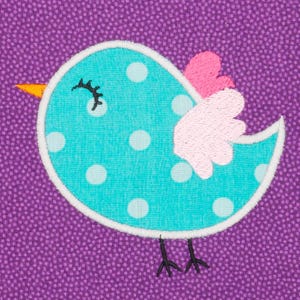 Puede incluir: Pájaro turquesa bordado con lunares blancos, pico amarillo y alas rosas y blancas. El pájaro está delineado en blanco y sobre un fondo morado punteado. El pájaro tiene patas negras y un ojo cerrado.
