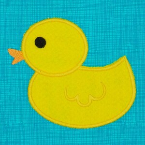 May include: A yellow felt rubber duck with a black eye and an orange beak. The duck is on a blue and white patterned background.