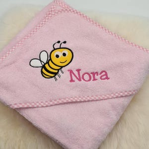May include: Pink hooded towel with a white and pink checkered trim. The towel features an embroidered bee with a yellow body and black stripes, and the name "Nora" is embroidered below the bee.