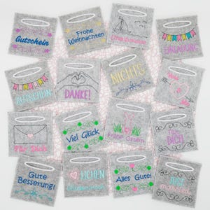 May include: A set of 14 grey felt gift tags with white halo embroidery. Each tag features a different embroidered design and text, including "Gutschein", "Frohe Weihnachten", "Liebe Grüße", "Einladung", "Danke", "Nichts", "You & Me", "Für Dich", "Viel Glück", "Frohe Ostern", "Gute Besserung", "Lichen Glückwunsch", "Alles Gute", and "Just Married".