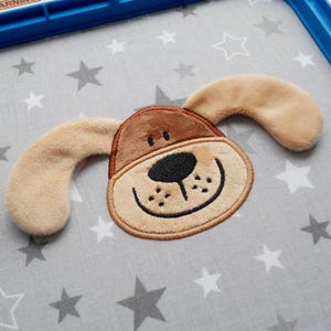 Puede incluir: Una cara de perro de peluche marrón con orejas caídas, bordada con una boca sonriente y una nariz negra. La cara del perro está sobre un fondo de tela gris con estrellas blancas.