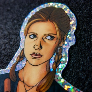 Close up of Buffy Summers sticker with glitter border and details. She is wearing a glittery cross and holding a wooden stake.