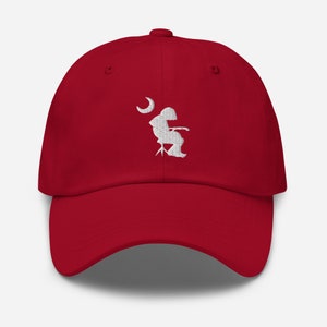 May include: A red baseball cap with a white embroidered design of a person sitting on a chair with a crescent moon above them.