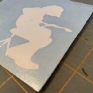 May include: White vinyl decal of a person sitting on a stool, playing a guitar. The decal is on a light blue background.