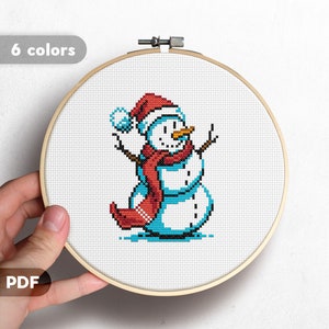 May include: Cross stitch pattern of a snowman wearing a red hat and scarf. The pattern is in a hoop and is ready to be stitched.