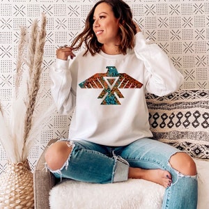 May include: White sweatshirt with a colorful eagle design. The eagle is a geometric design with a tribal pattern. The sweatshirt is worn by a person sitting on a couch.