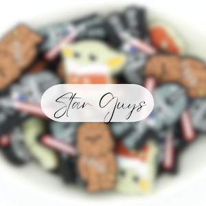 May include: A collection of colourful, cartoon-style iron-on patches featuring characters from the Star Wars franchise. The patches are arranged in a pile and include characters like Yoda, Darth Vader, and Stormtroopers. The text "Star Guys" is written on a white oval sticker.