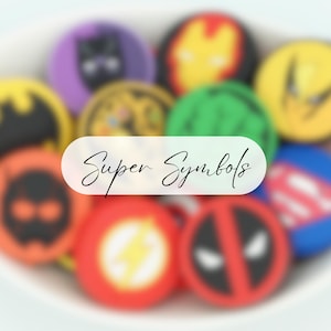 May include: A selection of vibrant, circular buttons featuring superhero emblems. The buttons showcase symbols and characters such as Batman, Iron Man, and Superman. The text "Super Symbols" is displayed on a white oval.