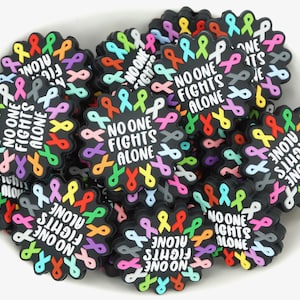 May include: A collection of black, flower-shaped charms with colorful ribbon designs. Each charm has the text "NO ONE FIGHTS ALONE" in white. The ribbons are in various colors, including pink, yellow, and blue.