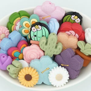 May include: A bowl filled with colorful silicone beads shaped like flowers, cacti, and other plants. The beads have a variety of colors, including pink, blue, green, yellow, and white.