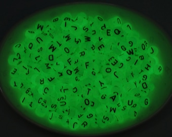May include: A close-up of a bowl filled with glow-in-the-dark letter beads. The beads are a bright green color and have black letters on them. The letters are a mix of uppercase and lowercase.