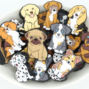 May include: A collection of colourful, cartoon dog charms in a white bowl. The charms feature different dog breeds, including a pug, a dalmatian, a golden retriever, and a dachshund.
