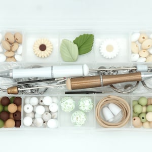 May include: A clear plastic storage box with compartments filled with various craft supplies, including beads, charms, and tools. The box contains white, brown, and green beads, as well as a white pen, a brown pen, and a metal tool.