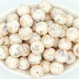 May include: A bowl of cream-coloured, round beads featuring illustrations of Winnie the Pooh characters. Each bead has a hole for threading. The illustrations are in soft, muted colours. The beads are in a white bowl.
