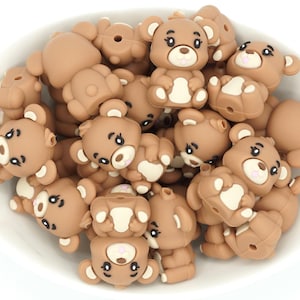 May include: A bowl filled with brown silicone baby teether beads shaped like teddy bears with white accents. The beads have black eyes and pink noses.