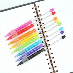 May include: A set of colourful mechanical pencils with different coloured tips. The pencils are arranged in a rainbow pattern on a notebook.