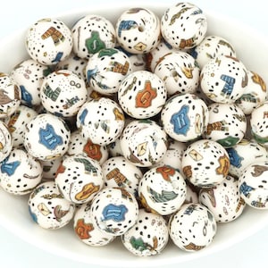 Peut inclure: Un ensemble de perles en céramique blanches avec des motifs colorés et fantaisistes. Chaque perle présente un motif unique de points et d'illustrations, notamment des chapeaux, des écharpes et d'autres éléments décoratifs. Les perles sont présentées dans un bol blanc.