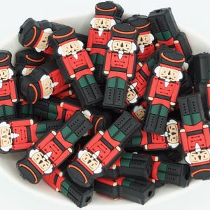 May include: A bowl filled with red, black, and green nutcracker figurines. The figurines feature a classic design with a black hat, red coat, and green trousers. The bowl is white and contains many figurines.