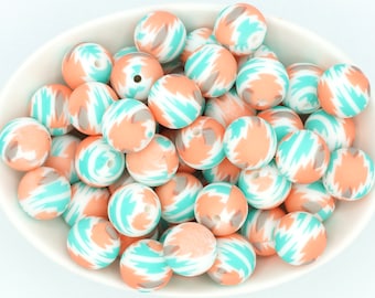 May include: A bowl of colorful silicone beads with a white, peach, and turquoise abstract pattern. The beads are round and have a hole in the center.