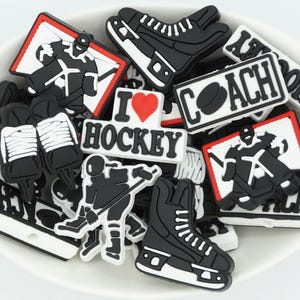 May include: A selection of black and white hockey-themed charms, featuring skates, players, and signs with "I ❤️ HOCKEY" and "COACH". The charms are in a white bowl.