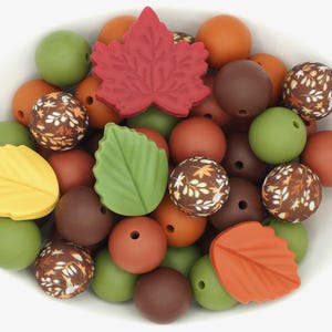 May include: A bowl filled with colourful silicone beads in various shapes, including leaves and maple leaves. The beads are in shades of brown, green, orange, and yellow.