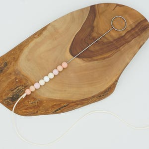 Puede incluir: Una tabla de madera con una cuerda de cuentas de silicona de color pastel enhebradas a trav&eacute;s de un bucle de metal. Las cuentas est&aacute;n dispuestas en un degradado de blanco a rosa.
