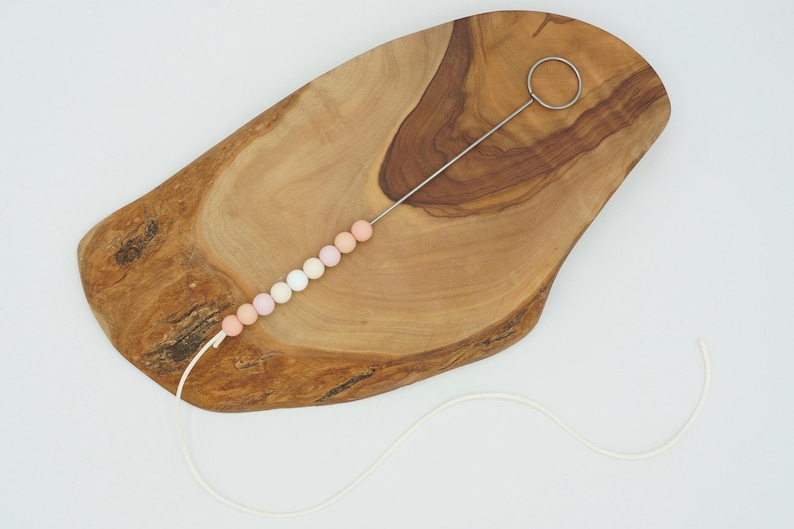 Puede incluir: Una tabla de madera con una cuerda de cuentas de silicona de color pastel enhebradas a trav&eacute;s de un bucle de metal. Las cuentas est&aacute;n dispuestas en un degradado de blanco a rosa.