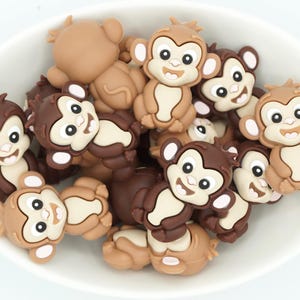 May include: A collection of monkey-shaped embellishments in various shades of brown, tan, and cream, arranged in a white bowl. Each monkey has a smiling face with black eyes and a cream-colored belly. The monkeys are made of a smooth, glossy material.