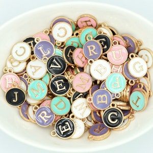 May include: A selection of colourful enamel alphabet charms in a white bowl. The charms have gold-coloured edges and are in various colours, including black, pink, purple, and teal. Each charm has a different letter.