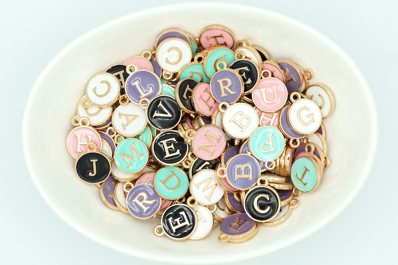 May include: A selection of colourful enamel alphabet charms in a white bowl. The charms have gold-coloured edges and are in various colours, including black, pink, purple, and teal. Each charm has a different letter.