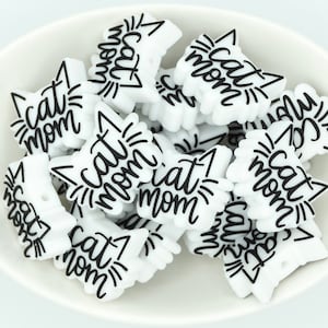 May include: A bowl filled with white silicone cat-shaped charms with black lettering that reads "Cat Mom".