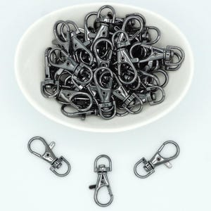May include: Gunmetal-coloured swivel snap hooks, presented in a white oval dish and scattered on a white surface. The metal hooks have a lobster claw clasp, ideal for various crafting projects.