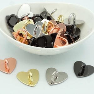 Pode incluir: Uma tigela cheia de ganchos de metal em forma de coração em vários acabamentos, incluindo prata, ouro, preto e ouro rosa. Os ganchos estão dispostos em uma pilha, com alguns sobrepostos. Há também quatro ganchos individuais sobre uma superfície branca.